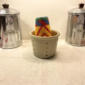 Könnte beinhalten: Eine weiße Keramik-Schale mit Löchern im Boden, in der ein gelbes, rotes und blaues Geschirrtuch liegt. Die Schale steht auf einer weißen Arbeitsplatte neben zwei silbernen Behältern mit den Aufschriften "Cafe" und "Sucre".