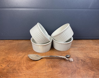 May include: Four white, ribbed ceramic ramekins are stacked on a wooden surface, with a silver spoon with a decorative handle. The ramekins are arranged in a group, and the spoon is placed in front of them. The background is a dark blue wall.