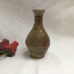 May include: A handmade ceramic vase with a narrow neck and a rounded body. The vase has a mottled brown and tan glaze with darker brown bands near the top. Decorative red roses are in the foreground.