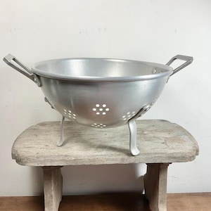 May include: A vintage aluminium colander with a wide bowl, two handles, and three legs. The colander has numerous small holes for draining and sits on a weathered wooden stool. The metal has a matte finish.