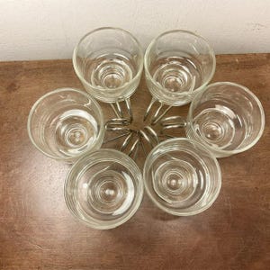 May include: Set of six clear glass cups with silver metal handles, arranged in a circle on a wooden surface.