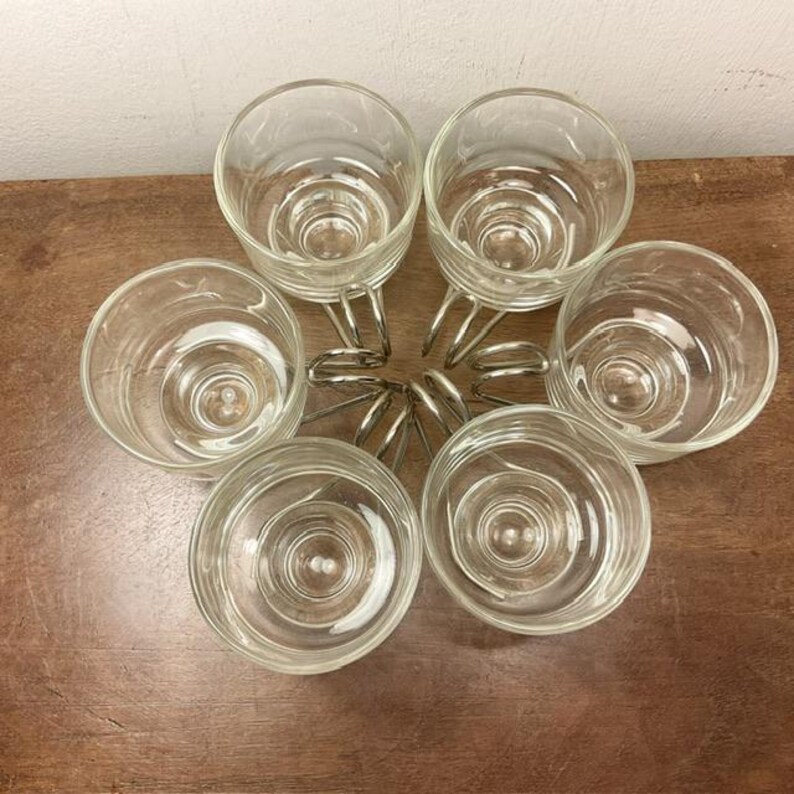 May include: Set of six clear glass cups with silver metal handles, arranged in a circle on a wooden surface.