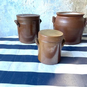 Pode incluir: Três potes de cerâmica esmaltada castanha com pegas laterais. Dois dos potes possuem tampas planas a condizer, enquanto o maior pote à direita está aberto. Cada recipiente apresenta detalhes estriados horizontais na borda superior.
