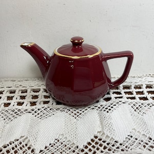 Op de afbeelding: Een diepbordeauxrode theepot met een gefacetteerd lichaam en een bijpassend deksel. De theepot is voorzien van een goudkleurige rand op het deksel en een goudkleurige tuit. Het heeft een stevig handvat en een gladde, glanzende afwerking.