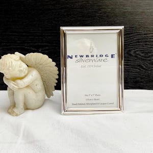 Peut inclure: Un cadre photo argenté et une petite statue de chérubin blanc cassé. Le cadre est rectangulaire et porte l'inscription "NEWBRIDGE Silverware" et peut contenir une photo de 13 cm x 18 cm. Le chérubin est assis, les ailes repliées.