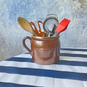 May include: A brown ceramic crock with a single handle containing various kitchen utensils, including a wooden spoon, orange plastic tools, a metal garlic press, and a bright red silicone basting brush, all standing upright inside the container.