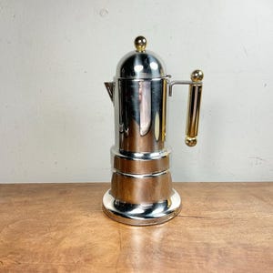 May include: A polished silver-tone metal stovetop espresso maker featuring a cylindrical body, a rounded lid with a small gold-tone knob, and a vertical handle with gold-tone accents at the top and bottom, reflecting light on its smooth surface.