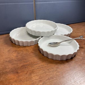 Op de afbeelding: Vier witte keramische taartschalen met geschulpte randen liggen op een houten oppervlak. Eén schaal bevat een zilveren lepel met een decoratief handvat. De schalen zijn leeg. De achtergrond bestaat uit donkerblauwe tegels.