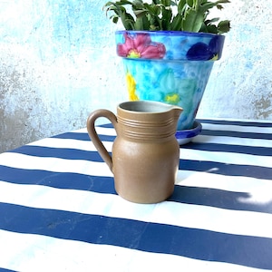 Pode incluir: Um jarro de grés castanho-claro com um acabamento mate suave, uma pega curva e três cristas horizontais à volta do gargalo. O jarro tem um corpo largo e arredondado e um pequeno bico, apresentando um design simples e funcional.