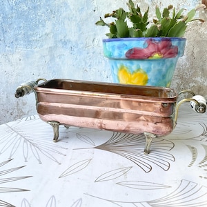 May include: A rectangular copper basin with a metallic finish, featuring four ornate silver-toned feet and two side handles with white ceramic accents. The vessel has a smooth, polished surface and a deep interior, designed with a classic clawfoot style.