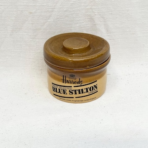 Könnte beinhalten: Ein zylindrischer Keramiktopf mit passendem Deckel, der eine glänzend braune Oberseite und eine hellbraune Basis aufweist. Die Seite zeigt schwarzen Text mit der Aufschrift Harrods, Finest Selected Blue Stilton Cheese und Harrods Limited Knightsbridge London England.