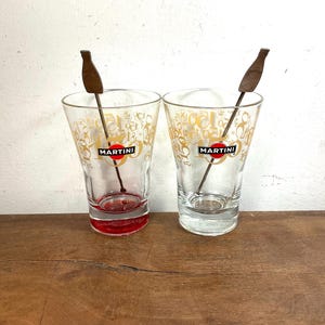 Op de afbeelding: Twee doorzichtige glazen met het Martini-logo in rood, zwart en wit, met goudkleurige cijfers op de buitenkant. Elk glas bevat een donkerbruine roerstaaf in de vorm van een fles. Eén glas heeft een rode onderkant.