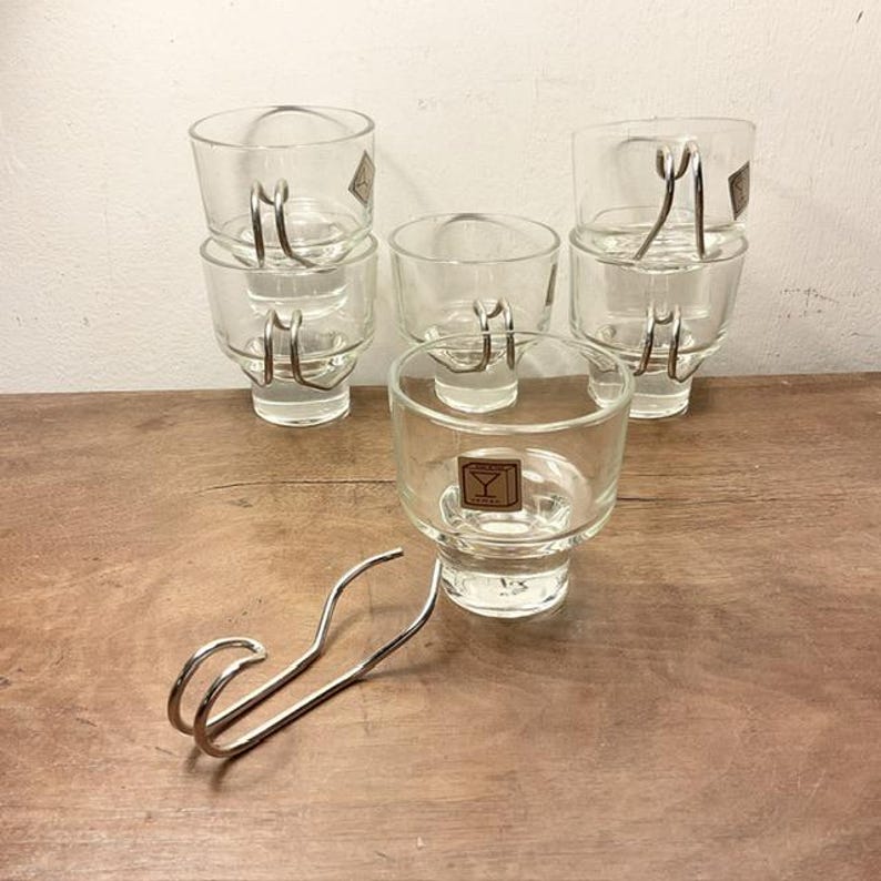May include: Set of six clear glass cups with metal handles. The cups are stacked in pairs and are arranged on a wooden surface. A metal hook is also visible in the image.