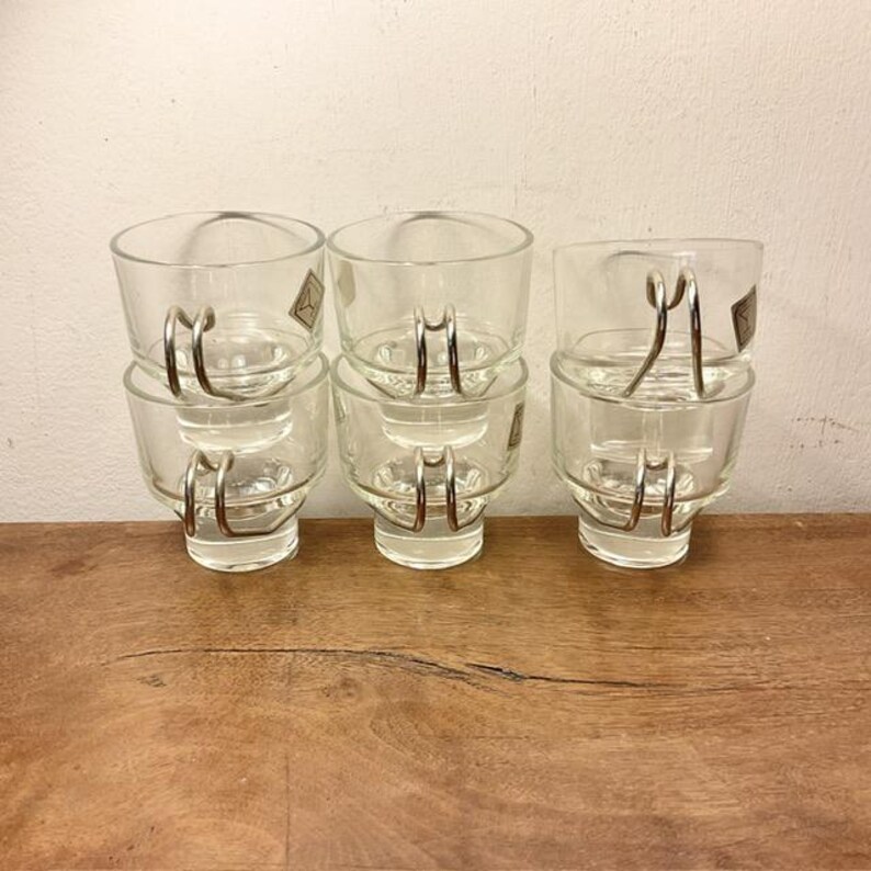 May include: Set of six clear glass cups with silver metal handles. The cups are stacked in pairs, with the handles facing outward.