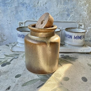 Puede incluir: Un tarro de cerámica marrón con un tapón de corcho. El tarro tiene un cuerpo redondeado y un borde ligeramente ensanchado. El fondo incluye recipientes de esmalte blanco con texto azul y una bandeja blanca.