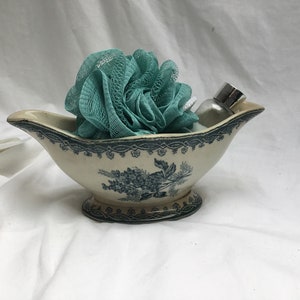 May include: A white ceramic gravy boat with a blue floral design, filled with a teal bath pouf and a clear glass bottle with a silver lid.