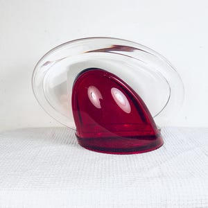 May include: A clear glass sculpture with a deep red, teardrop-shaped element. The clear glass forms a curved, oval shape around the red piece. The sculpture sits on a white surface, creating a striking contrast.