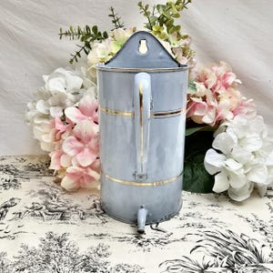 Könnte beinhalten: Eine alte, hellblaue Emaille-Kaffeekanne mit goldenen Verzierungen. Die Kanne hat einen Griff und einen Ausguss. Der Hintergrund zeigt rosa und weiße Blumen.
