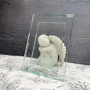 May include: A clear glass photo frame with a small, white cherub statue inside. The cherub is in a seated position with its wings spread. The frame is angled, and the background is a dark wood surface.