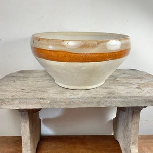 Op de afbeelding: Een crèmekleurige keramische kom met een brede, oranjebruine band in het midden. De kom staat op een verweerde, lichtgrijze houten kruk met drie poten. De kom heeft een brede opening en een afgeronde vorm.