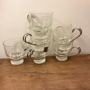May include: Set of six clear glass cups with metal handles. The cups are stacked on top of each other.