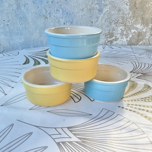 Puede incluir: Cuatro ramequines de cerámica en colores pastel: dos azules claros y dos amarillos. Los ramequines están apilados y colocados sobre un mantel blanco con un diseño floral. El fondo es una pared azul claro.