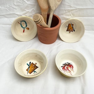 May include: Four small white bowls with yellow rims, each featuring a different kitchen utensil illustration. The bowls are arranged around a terracotta pot with the word "Kitchen" written on it.
