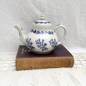 Puede incluir: Una tetera de cerámica blanca con tapa, adornada con diseños florales azules. La tetera está sobre un libro antiguo con una cubierta marrón desgastada. El fondo es una superficie blanca texturizada.