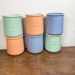 Puede incluir: Seis tarros de cerámica en colores pastel: verde menta, melocotón y azul claro. Cada tarro tiene forma cilíndrica con interior blanco y la palabra "FERMIERE" en relieve en el frente. Algunos tarros están apilados.