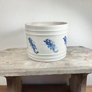 Puede incluir: Un tarro de cerámica blanco con diseños florales azules. El recipiente cilíndrico tiene estrías horizontales y se exhibe sobre un taburete de madera desgastada. El patrón floral azul se repite alrededor del exterior del tarro.