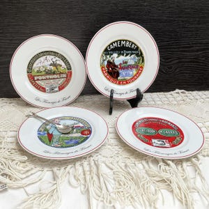 Può includere: Quattro piatti in ceramica bianca con bordi rossi, con etichette di formaggio francesi vintage. I piatti presentano illustrazioni colorate e testo, tra cui "Camembert" e "Les Fromages de France". Un piccolo cucchiaio d'argento è su un piatto.