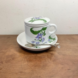 Könnte beinhalten: Eine weiße Keramik-Teetasse mit passendem Deckel, Untertasse und Löffel. Die Tasse und der Deckel weisen ein Blumendesign mit lila Blüten und grünen Blättern auf. Der Löffel hat ein dekoratives Kristallende.