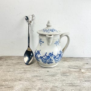 Puede incluir: Una crema de cerámica blanca con un diseño floral azul y una tapa a juego, junto a una cuchara plateada con un mango decorativo. La cuchara y la crema están sobre una superficie de madera.