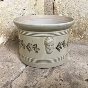 May include: A white ceramic planter with a brown floral design and a face in relief. The planter has a round shape and a wide rim.