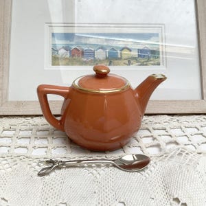 May include: A small, orange ceramic teapot with a gold trim and a matching lid. A silver spoon with a decorative handle is in front of the teapot. A framed picture of colorful beach huts is in the background.
