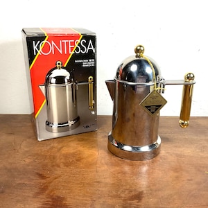 Pode incluir: Uma cafeteira de aço inoxidável prateado com alça e botão da tampa dourados. A cafeteira apresenta um corpo cilíndrico, um bico e uma etiqueta em forma de losango presa por um cordão amarelo, ao lado da caixa original rotulada KONTESSA.
