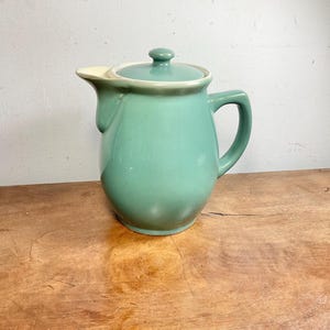 以下が含まれることがあります： 滑らかでマットな質感のセージグリーンの陶器製ティーポット。丸みを帯びた本体、右側に湾曲したハンドル、左側に短い注ぎ口があり、上部中央に小さなつまみ付きの蓋が付属しています。