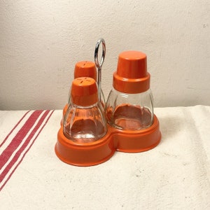 Pode incluir: Conjunto de condimentos de vidro e plástico laranja com três recipientes e uma grelha de metal. Os recipientes têm corpos de vidro transparente e tampas de plástico laranja.