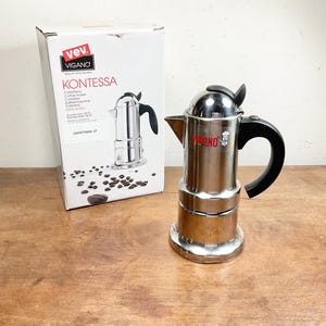 May include: A stainless steel Vev Vigano Kontessa stovetop coffee maker with a black handle and lid knob. The coffee maker is positioned next to its white product box, which features the brand name, the model name, and an image of the coffee maker.