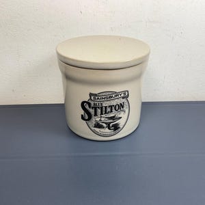 Puede incluir: Un tarro de cerámica beige con tapa a juego. La parte frontal presenta una etiqueta impresa en negro que dice Sainsbury's Blue Stilton, acompañada de una ilustración de una rueda de queso sobre un pedestal y texto pequeño abajo.