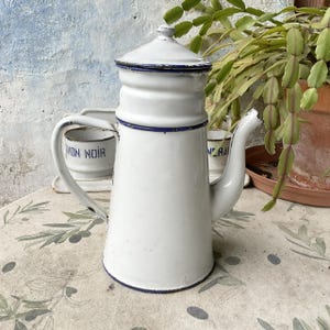 Könnte beinhalten: Weiße Emaille-Kaffeekanne mit blauem Rand und Deckel. Die Kanne hat einen langen Ausguss und einen Griff. Die Worte "WON NOIR" sind auf einer kleinen Tasse im Hintergrund zu sehen. Die Kanne steht auf einem Tisch mit floralem Muster.