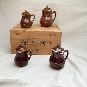 May include: Four brown ceramic creamer pitchers with white lids. The pitchers are arranged on a wooden box with the text "Chateau Margaux du Roi Saint-Estèphe 2006" printed on the side.