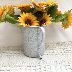 Op de afbeelding: Een witte gespikkelde metalen kan met een handvat, gevuld met een boeket gele zonnebloemen.