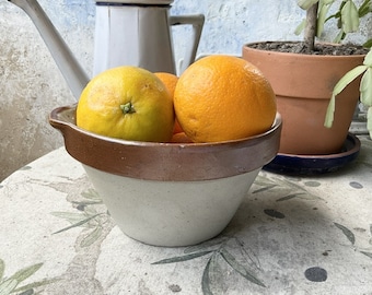 May include: A white ceramic bowl with a brown rim, filled with three oranges. The bowl is sitting on a table with a floral pattern.