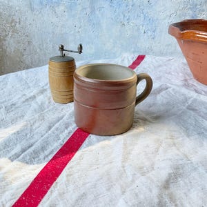 May include: A brown ceramic mug with a cylindrical body and a side handle, positioned next to a small wooden barrel-shaped pepper mill and a large terracotta bowl on a white fabric surface with a prominent red stripe running across the centre.