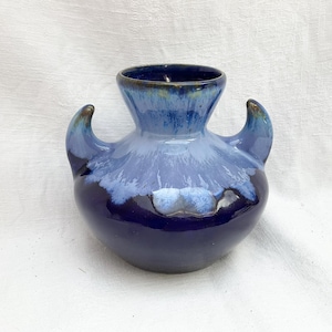 Op de afbeelding: Een keramische vaas met een rond lichaam en twee gebogen handvatten. Het glazuur verloopt van een diep, donker marineblauw aan de basis naar een lichter, gevlekt hemelsblauw bij de hals en rand, wat een opvallend druipeffectpatroon creëert.