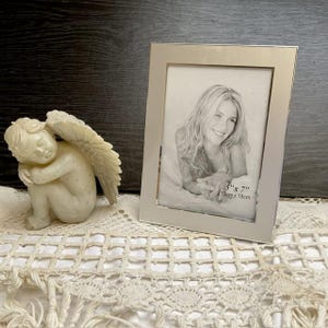 Op de afbeelding: Een zilveren fotolijst met een witte passe-partout en een foto van een vrouw erin. De lijst is 5 inch bij 7 inch en meet 13 cm bij 18 cm.