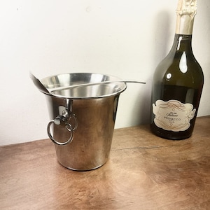Puede incluir: Un cubo de hielo de plata con una cuchara a juego, utilizado para enfriar bebidas. El cubo tiene dos anillos en el lateral. Una botella de prosecco está en el fondo.