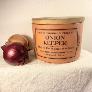 May include: An orange ceramic onion keeper with a wooden lid, labeled "The Original Suffolk Onion Keeper." A red onion and a brown onion sit beside the container. The text on the keeper reads "Ideal for Use at Home or on Safari."