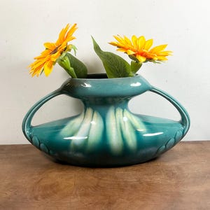 Peut inclure: Un vase en céramique turquoise avec deux anses, contenant deux tournesols jaunes avec des feuilles vertes. Le vase a une forme unique avec une base large et un sommet plus étroit. Le vase est sur une surface en bois brun.